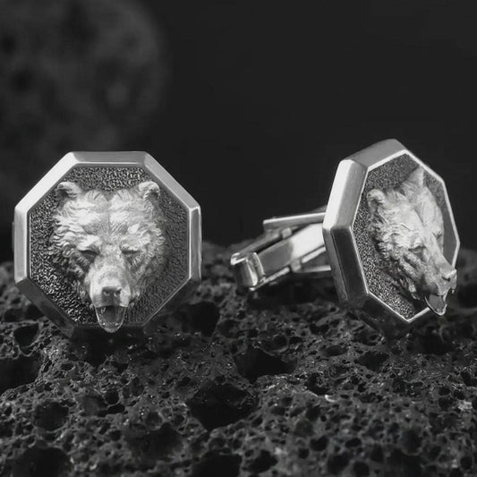 An oxidized bear head cufflink pair rests on dark volcanic rock in a macro studio close-up, showing a sculpted grizzly-inspired animal face inside octagon frames.