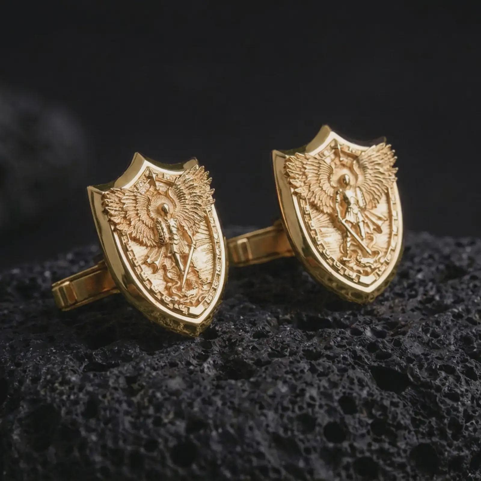 Gold plated Saint Michael cufflinks sit on textured volcanic rock in a dramatic studio macro shot, highlighting the Catholic archangel protection motif.