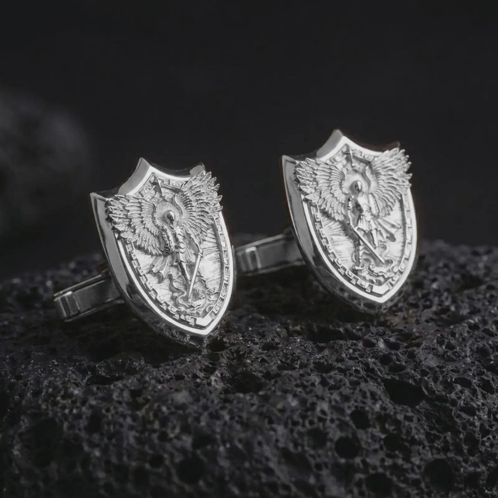 Polished silver Saint Michael cufflinks are displayed on black volcanic rock in a premium macro studio close-up with sharp shield edges and sacred warrior detail.