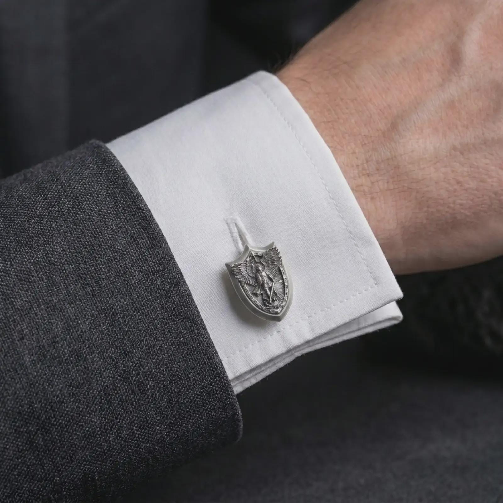 An oxidized Saint Michael cufflink is worn on a white shirt cuff in a close-up lifestyle shot, styled with a dark suit as a meaningful Catholic accessory.