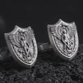 Load image into Gallery viewer, An oxidized Saint Michael cufflink pair rests on dark volcanic rock in a macro studio close-up, showing a shield-shaped archangel warrior design with detailed wings.
