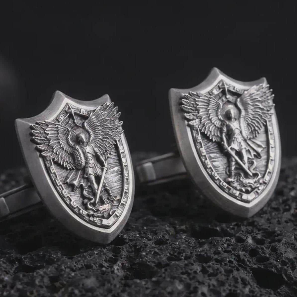An oxidized Saint Michael cufflink pair rests on dark volcanic rock in a macro studio close-up, showing a shield-shaped archangel warrior design with detailed wings.