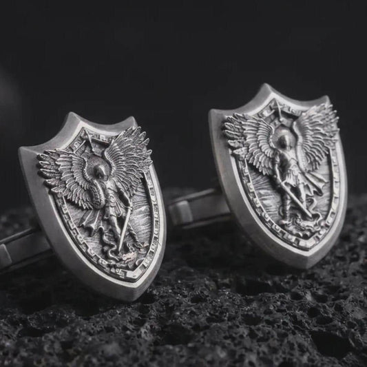 An oxidized Saint Michael cufflink pair rests on dark volcanic rock in a macro studio close-up, showing a shield-shaped archangel warrior design with detailed wings.