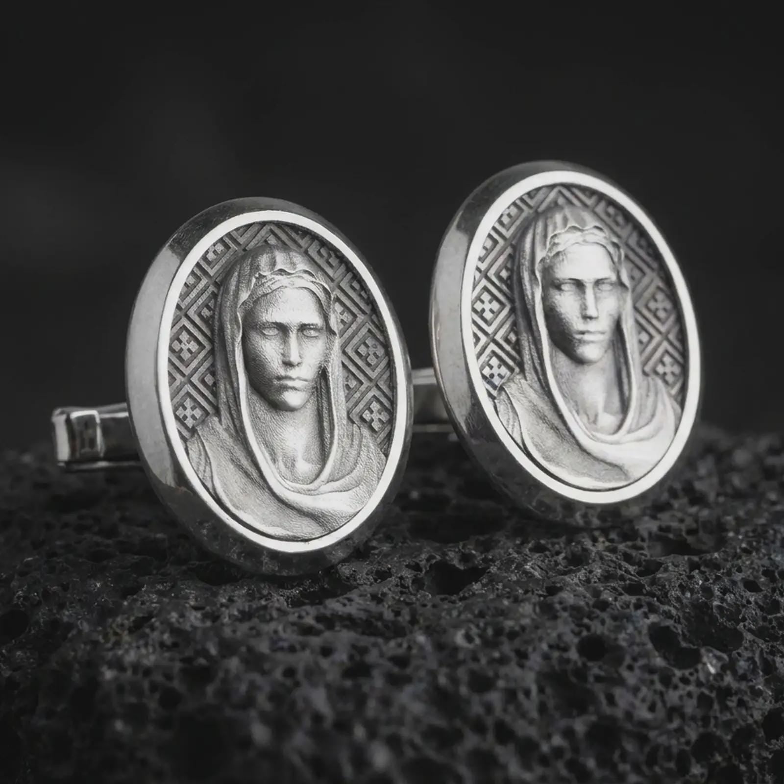 An oxidized Virgin Mary cufflink pair rests on dark volcanic rock in a macro studio close-up, showing a refined Marian portrait inside oval frames.