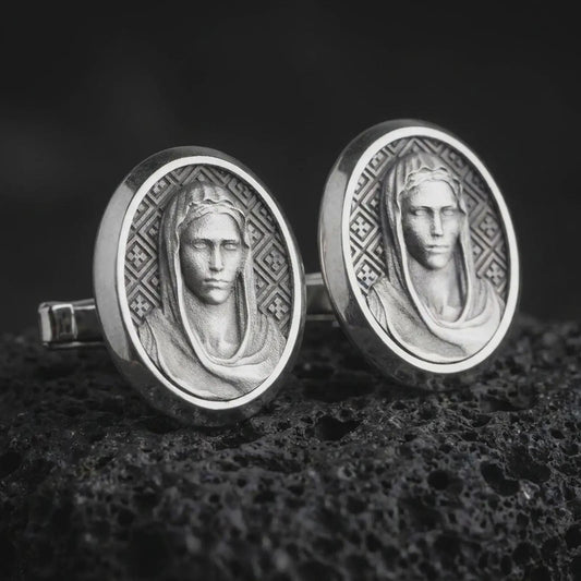 An oxidized Virgin Mary cufflink pair rests on dark volcanic rock in a macro studio close-up, showing a refined Marian portrait inside oval frames.
