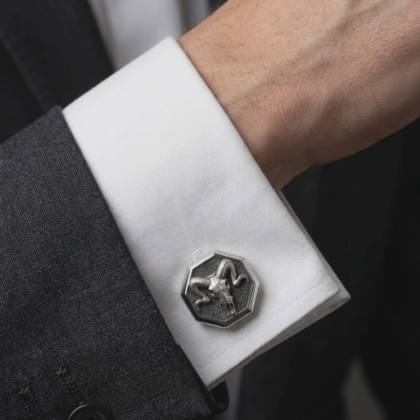 An oxidized ram skull cufflink is worn on a white shirt cuff in a close-up lifestyle shot, styled with a dark suit for a bold gothic formal look.
