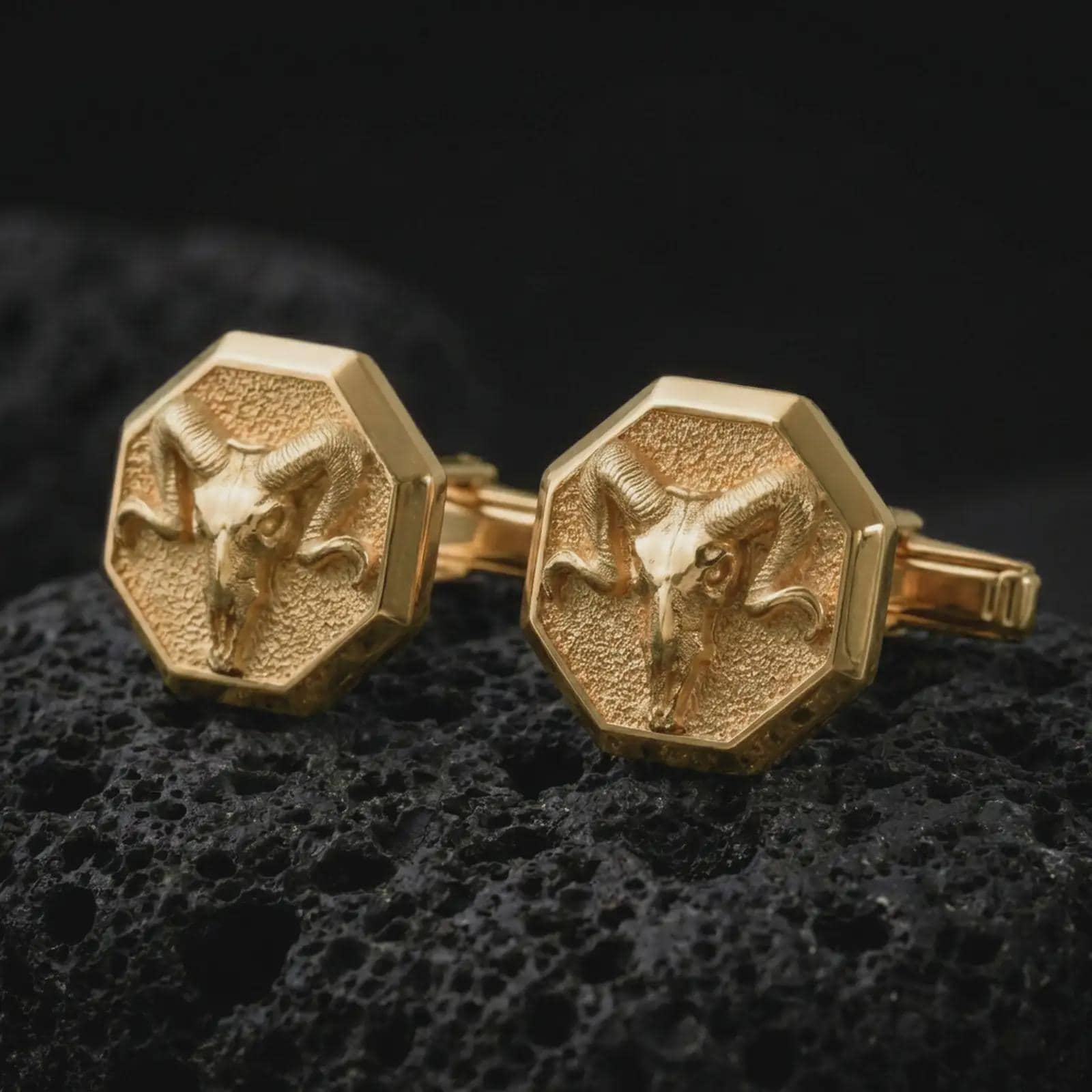 Gold plated ram skull cufflinks sit on textured volcanic rock in a dramatic studio macro shot, highlighting the Aries-inspired skull motif and formal accessory design.