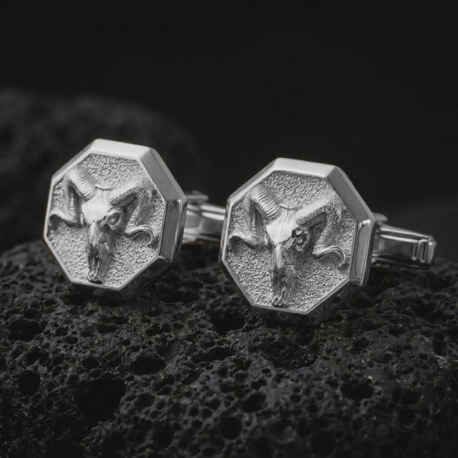 Polished silver ram skull cufflinks are displayed on black volcanic rock in a premium macro studio close-up with sharp horn texture and clean geometric edges.