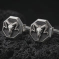 Load image into Gallery viewer, An oxidized ram skull cufflink pair rests on dark volcanic rock in a macro studio close-up, showing curved horns and sculpted skull detail inside octagon frames.
