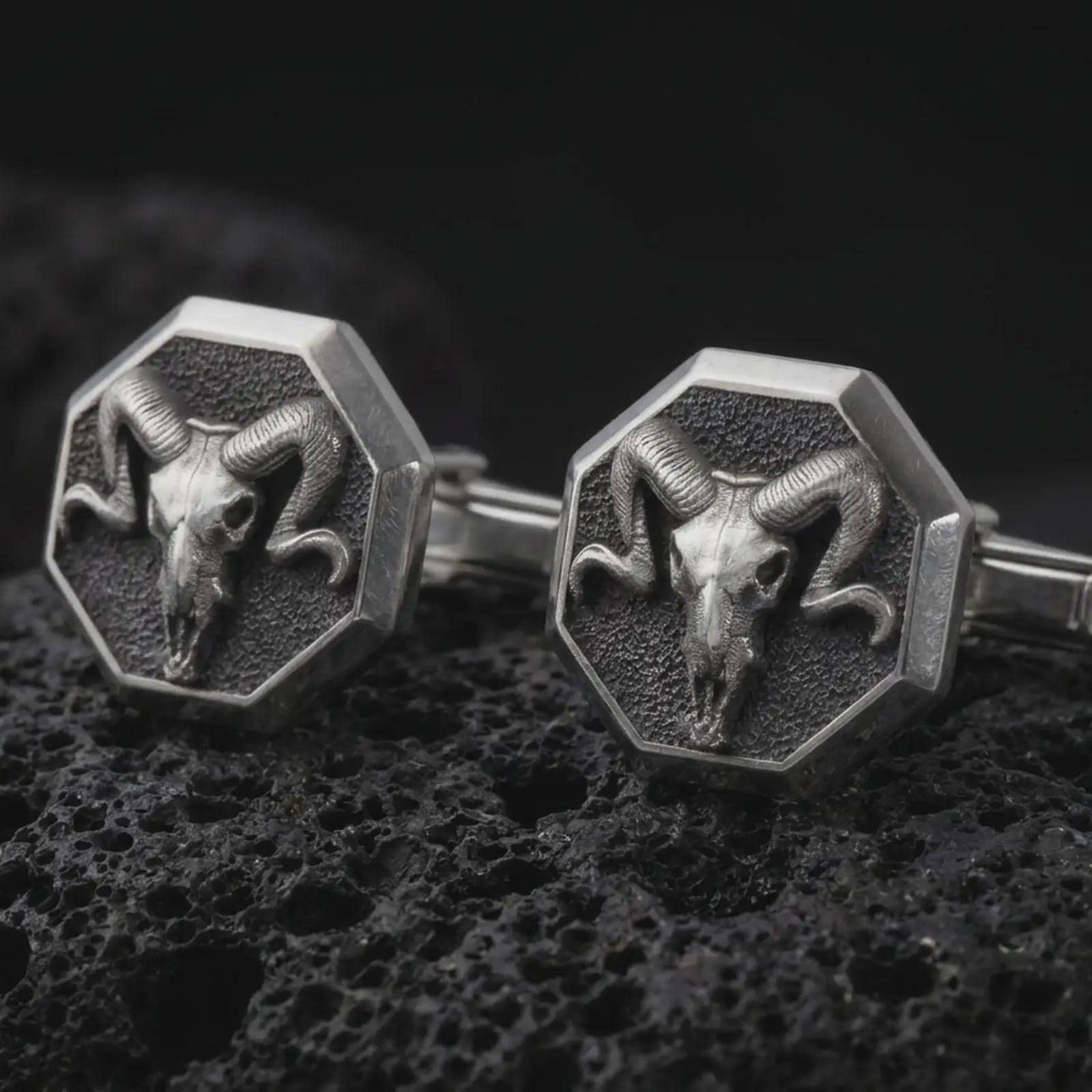 An oxidized ram skull cufflink pair rests on dark volcanic rock in a macro studio close-up, showing curved horns and sculpted skull detail inside octagon frames.