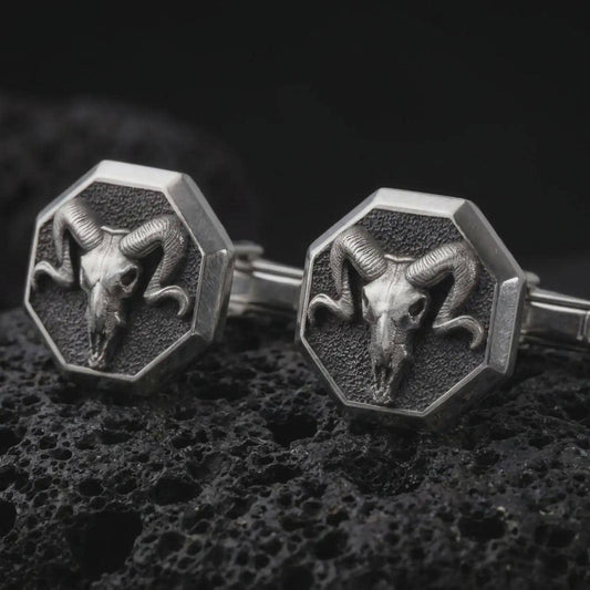 An oxidized ram skull cufflink pair rests on dark volcanic rock in a macro studio close-up, showing curved horns and sculpted skull detail inside octagon frames.
