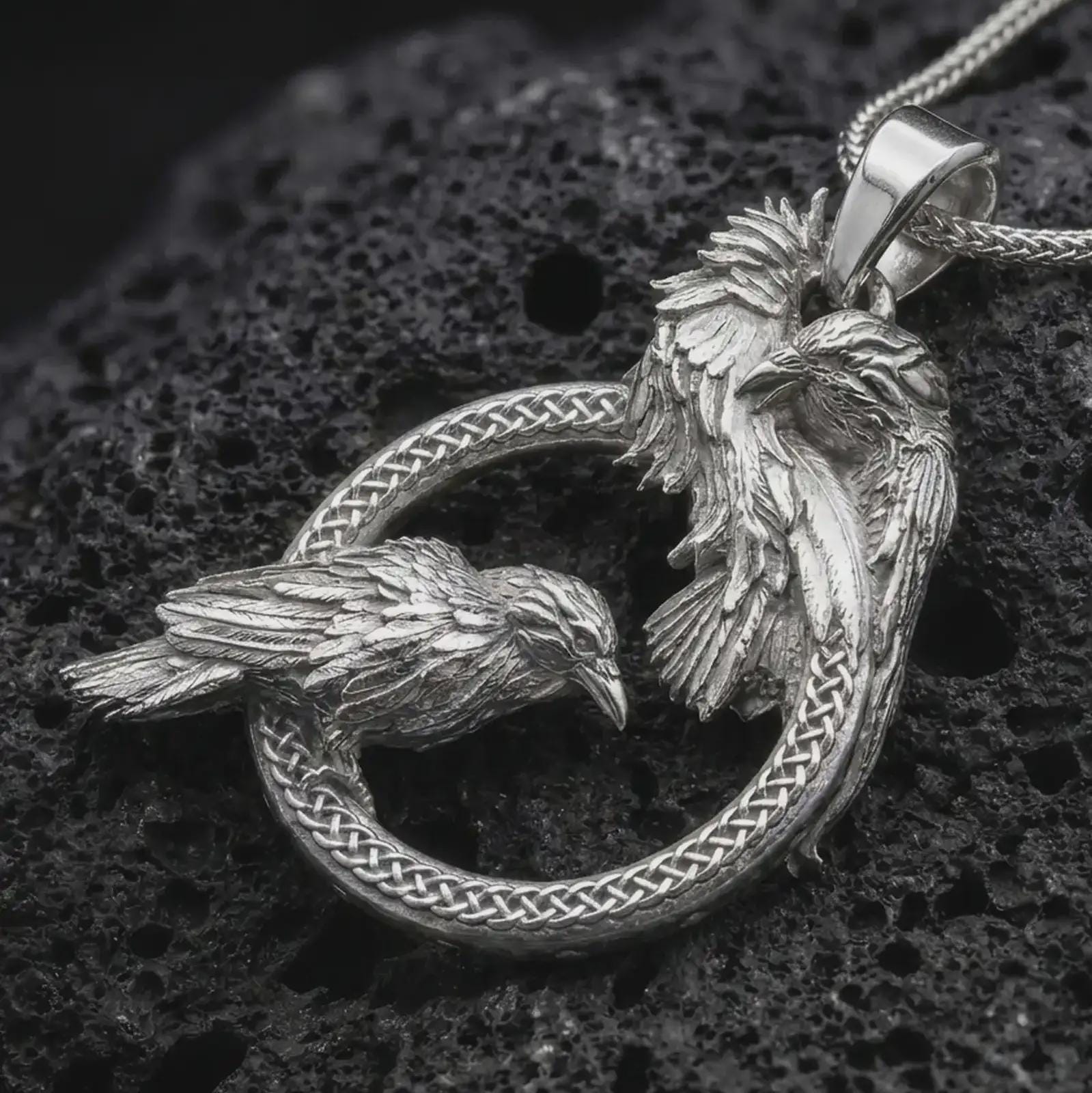Polished silver Hugin and Munin ravens pendant necklace on textured volcanic rock, centered macro jewelry photo with detailed Norse bird relief and bright silver highlights.