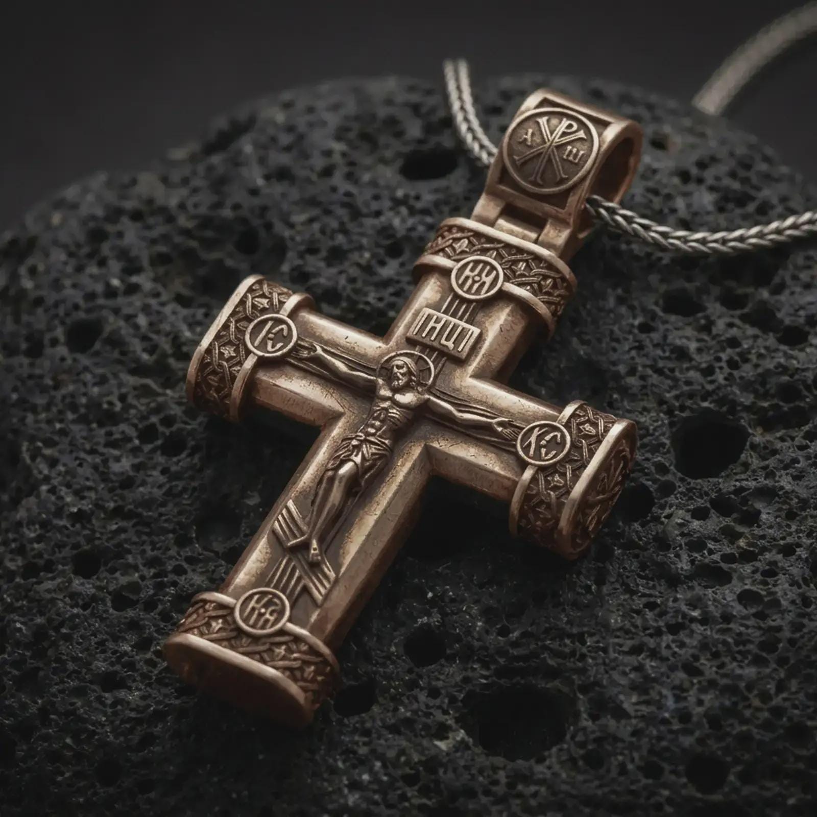Bronze crucifix cross pendant necklace on textured volcanic rock, close-up studio shot highlighting the Jesus figure, INRI plaque, ornate barrel ends, and antique silver contrast.