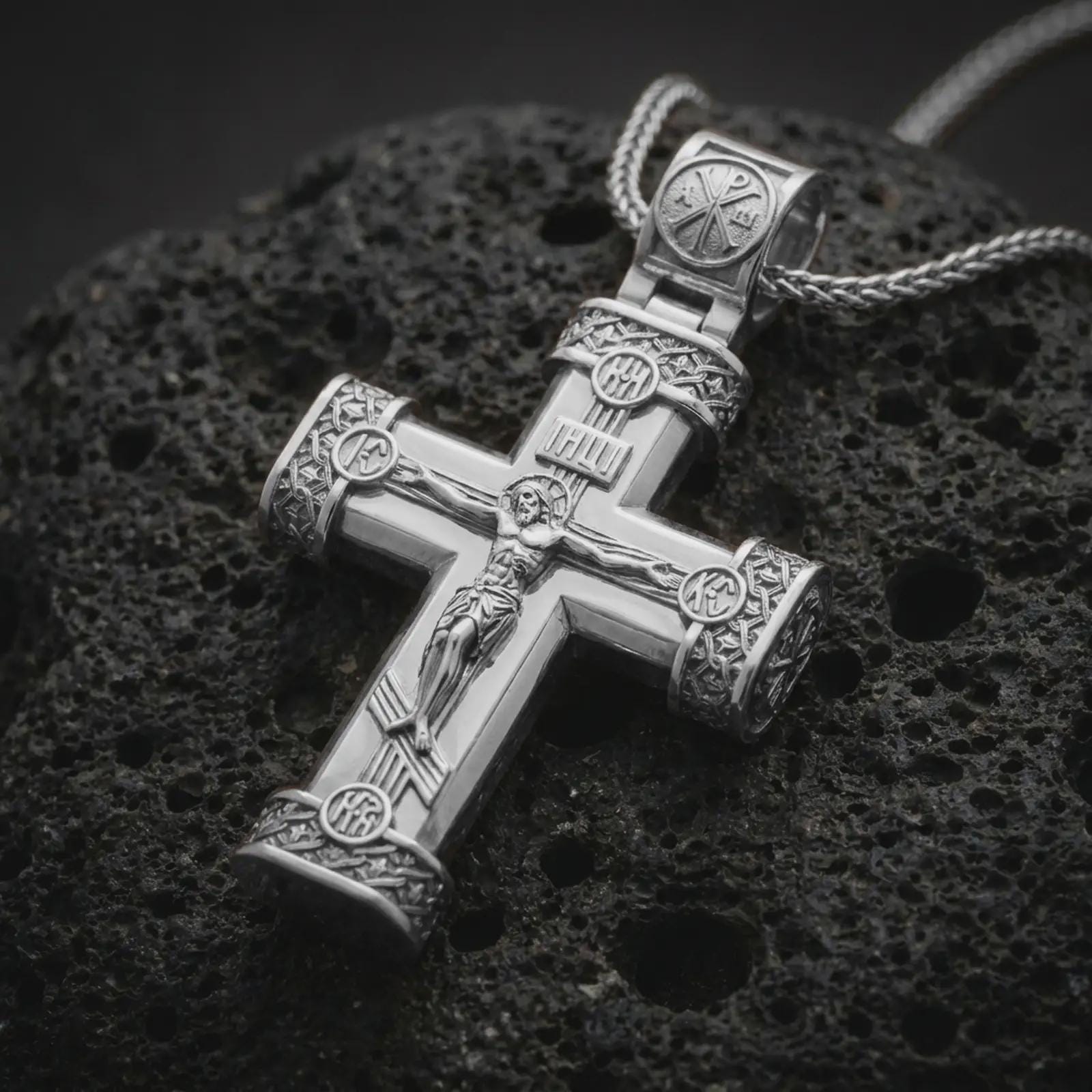 Polished silver crucifix cross pendant necklace on textured volcanic rock, centered macro jewelry photo with detailed Christ relief, engraved accents, and bright silver highlights.