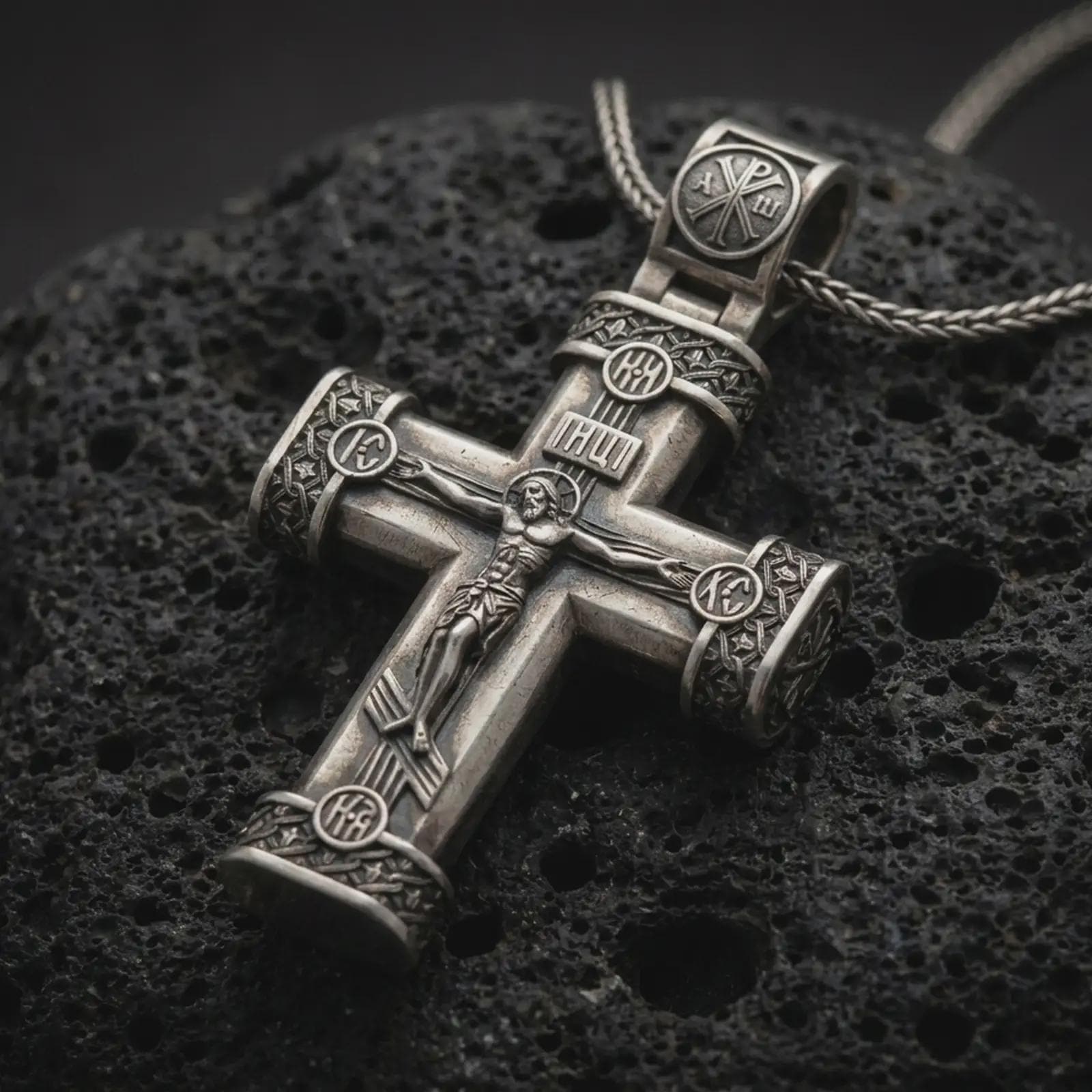 Oxidized crucifix cross pendant necklace on textured volcanic rock, close-up studio shot highlighting the Jesus figure, INRI plaque, ornate barrel ends, and antique silver contrast.