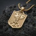 Load image into Gallery viewer, Gold plated German eagle shield pendant necklace on textured volcanic rock, premium close-up studio composition emphasizing the national eagle symbol, shield silhouette, and bold heraldic design.

