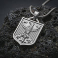 Load image into Gallery viewer, Polished silver German eagle shield pendant necklace on textured volcanic rock, centered macro jewelry photo with detailed bird crest, vertical wing lines, and bright silver highlights.
