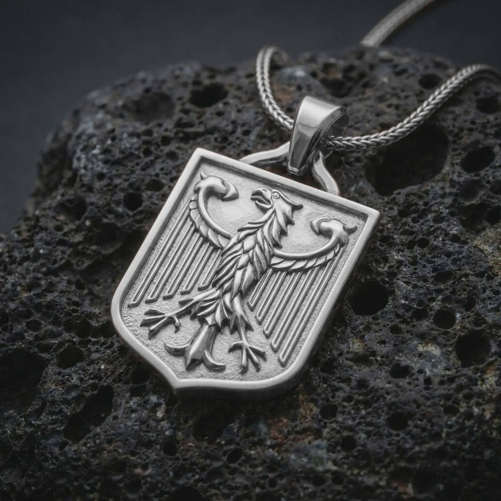Polished silver German eagle shield pendant necklace on textured volcanic rock, centered macro jewelry photo with detailed bird crest, vertical wing lines, and bright silver highlights.