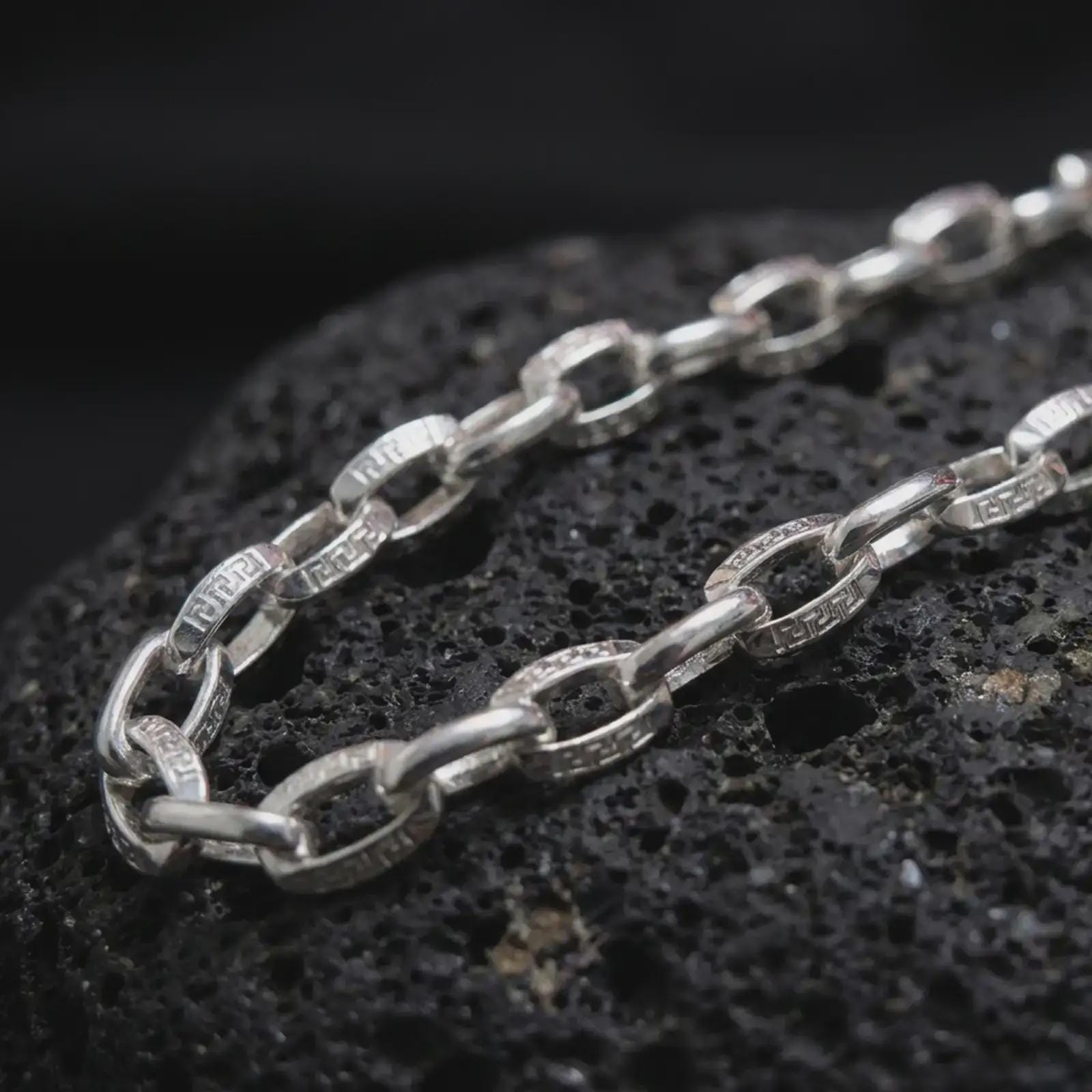 Polished silver Greek key chain necklace is displayed on black volcanic rock in a premium macro studio close-up with clean link structure and geometric accents.