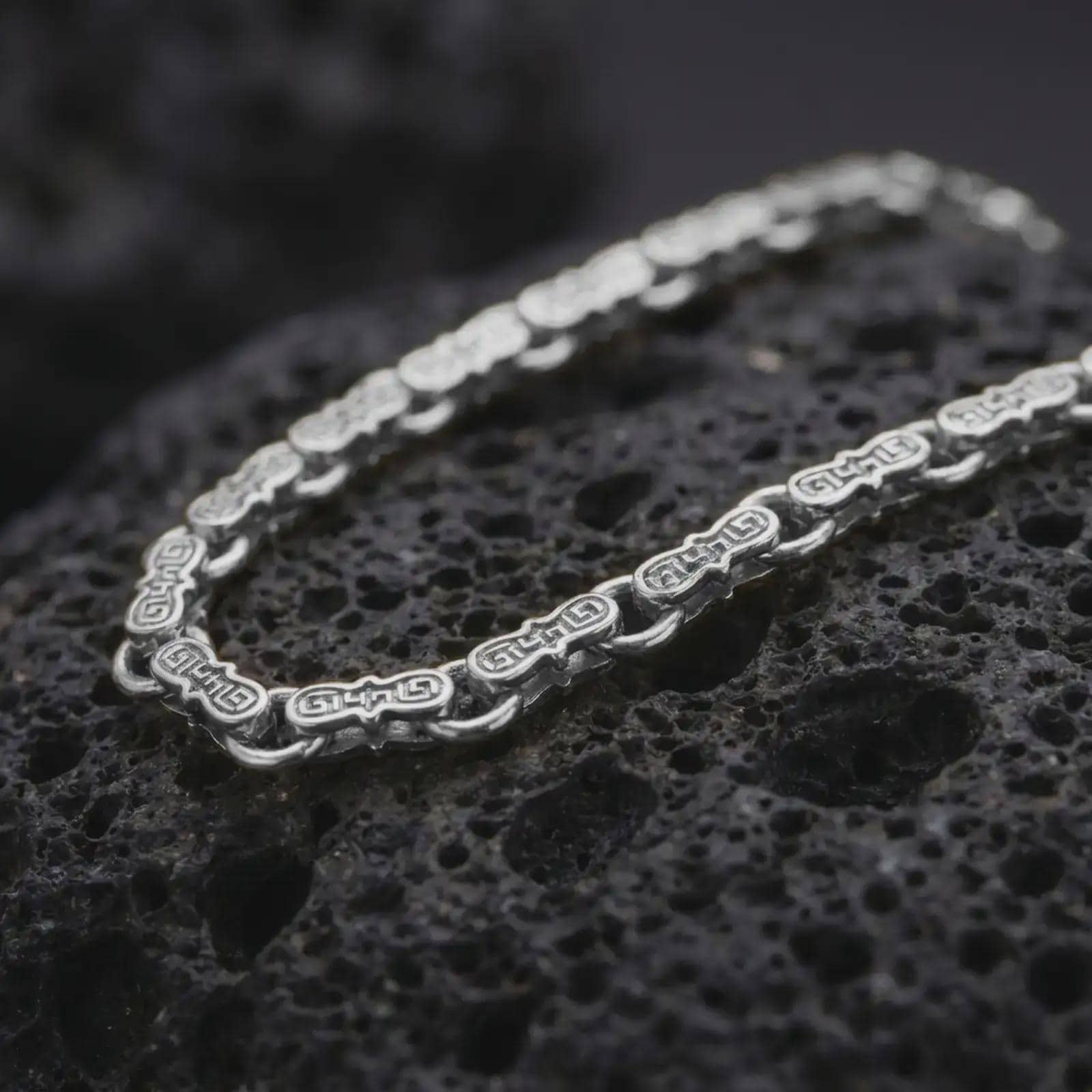 Polished silver Greek key chain necklace is displayed on black volcanic rock in a premium macro studio close-up with bright geometric link details.