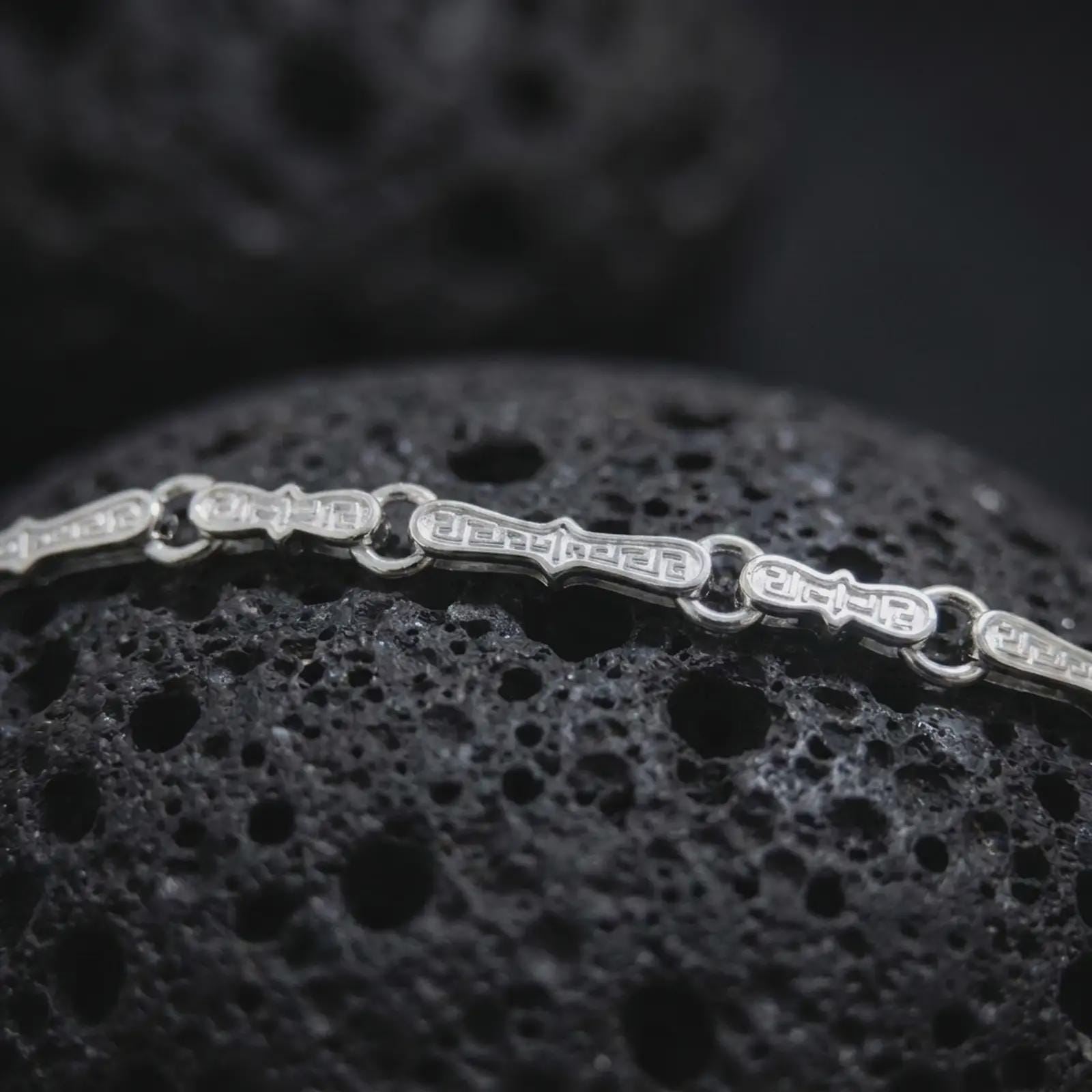 Polished silver Greek key chain necklace is displayed on black volcanic rock in a premium macro studio close-up with clean geometric link details.