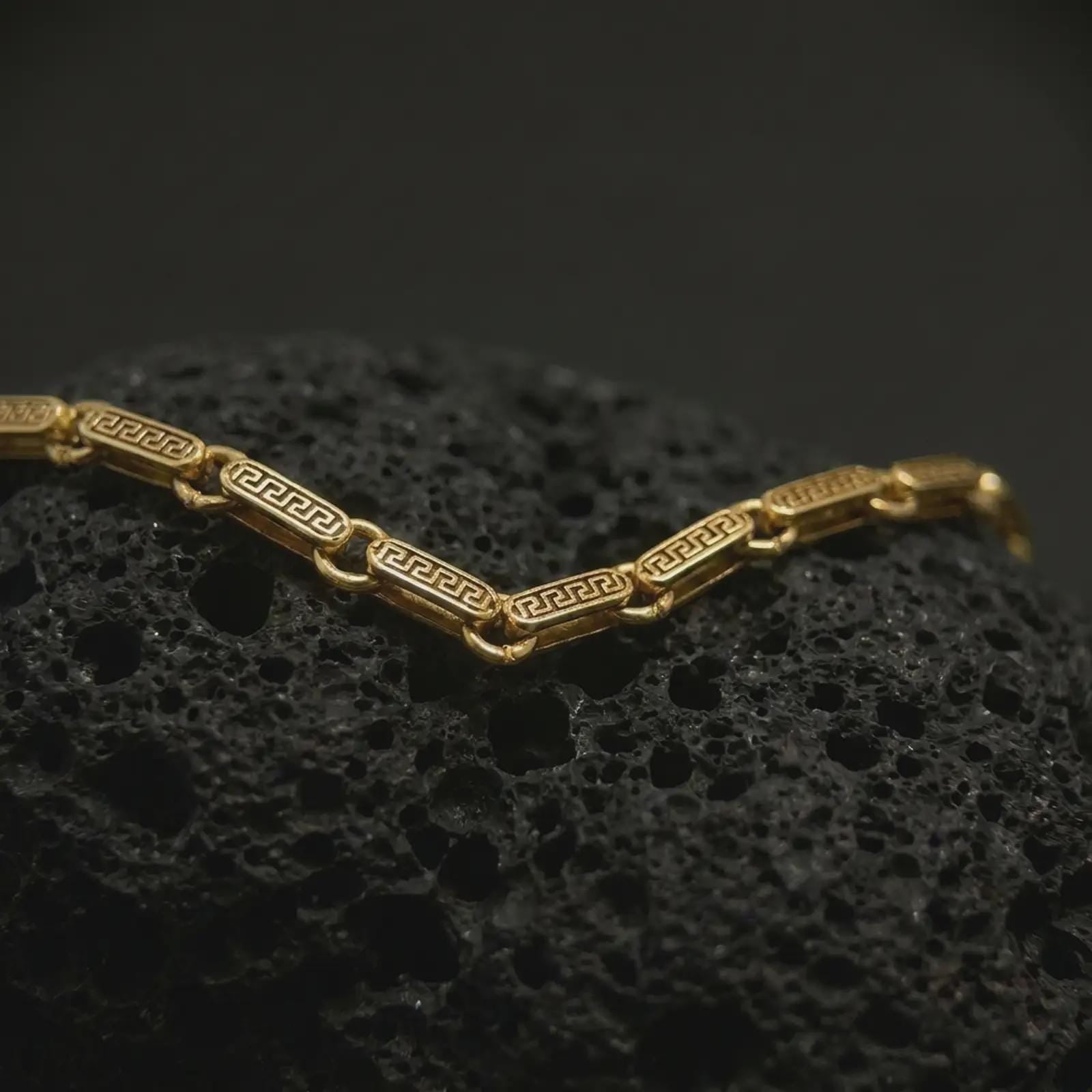 Gold plated Greek key chain laid across textured volcanic rock, emphasizing the classical meander motif and structured link-by-link construction.