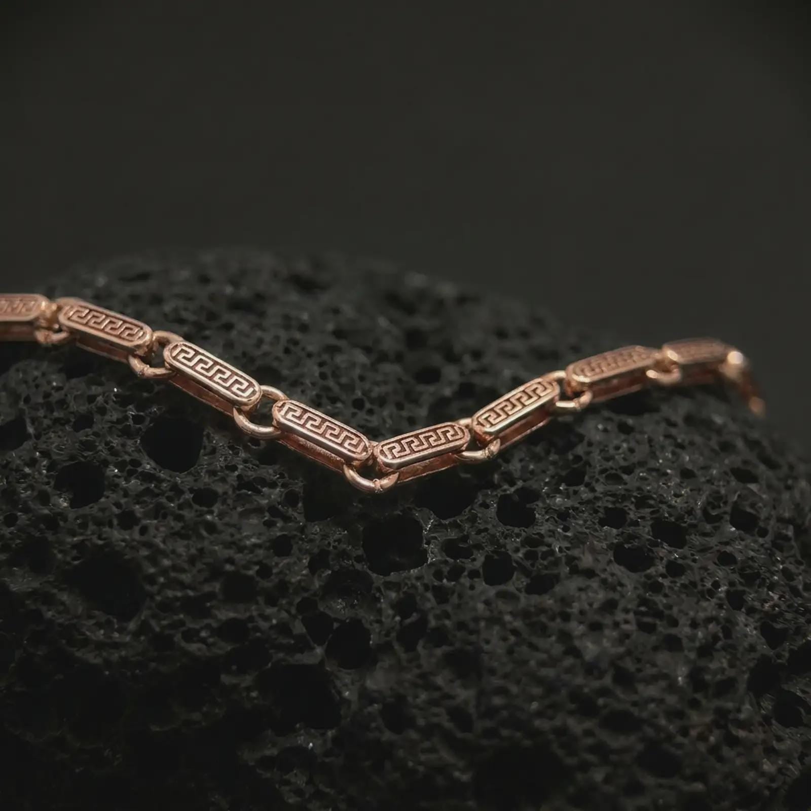 Rose gold plated Greek key chain photographed on black volcanic rock, showing its decorative patterned links and ancient Greek inspired design.