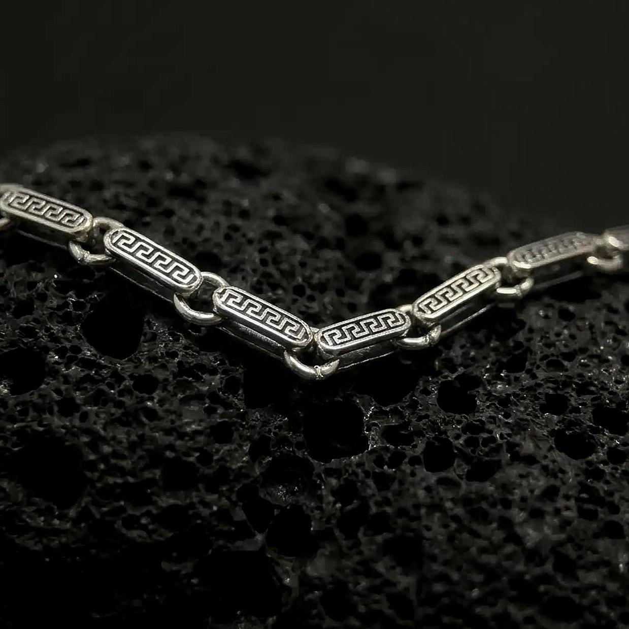 Oxidized sterling silver Greek key chain shown across black volcanic rock, highlighting the antique meander pattern engraved into each rectangular link.