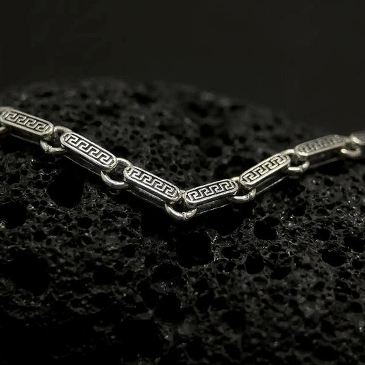 Oxidized sterling silver Greek key chain shown across black volcanic rock, highlighting the antique meander pattern engraved into each rectangular link.