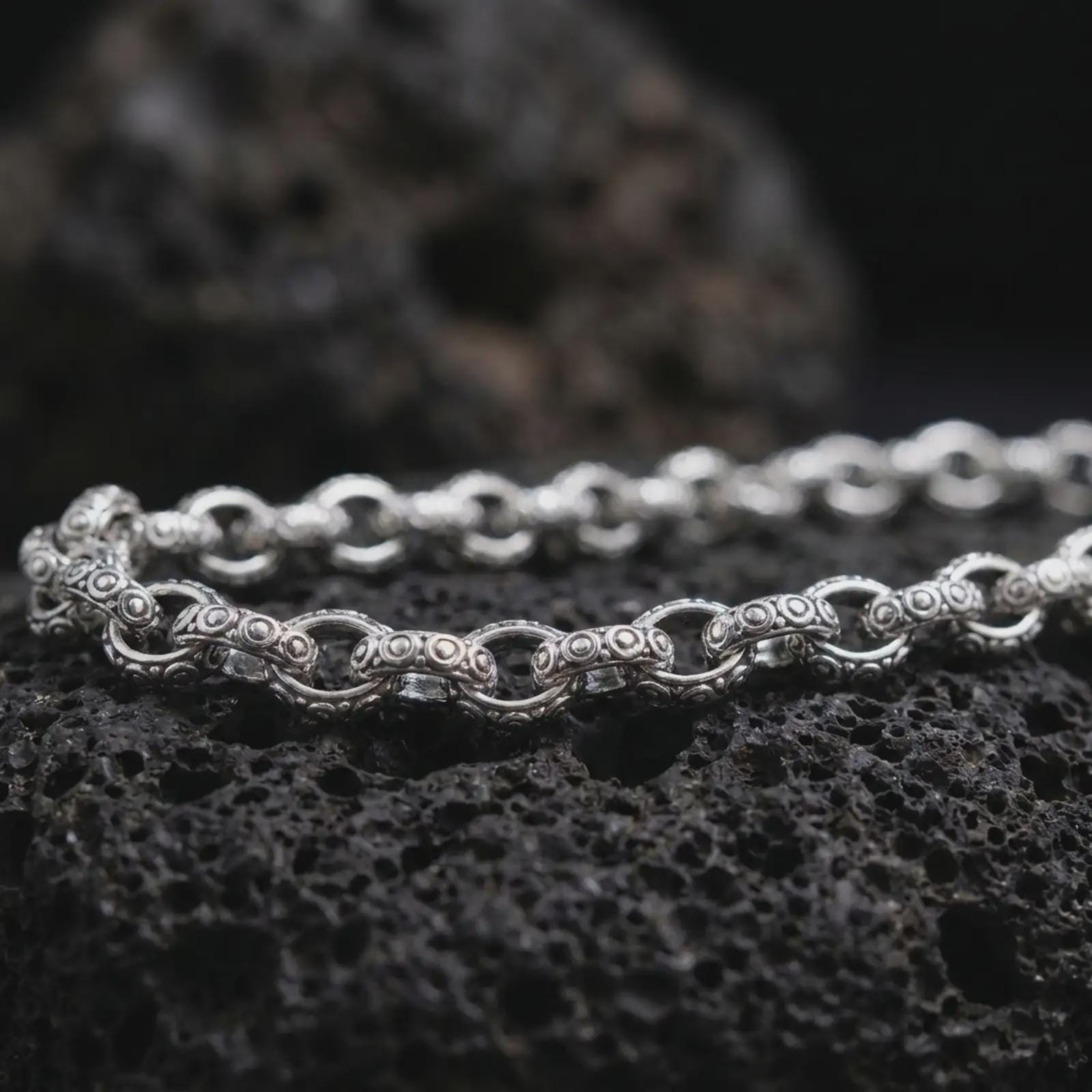 Polished silver ornamental chain necklace on volcanic stone, highlighting the sculpted link structure and bright reflective edges.