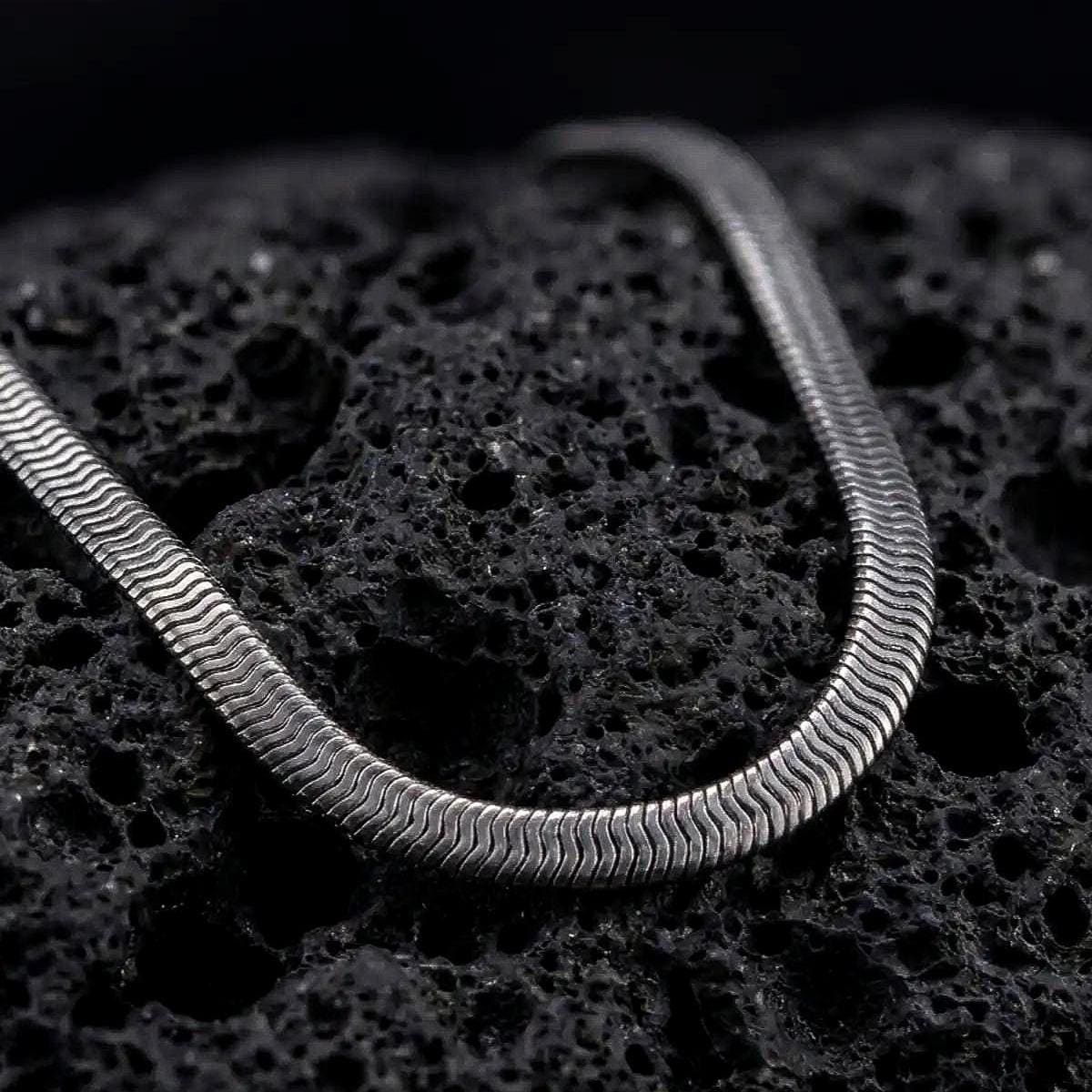 Oxidized sterling silver snake chain displayed on black volcanic rock, showing its smooth rounded profile and close flexible link structure.