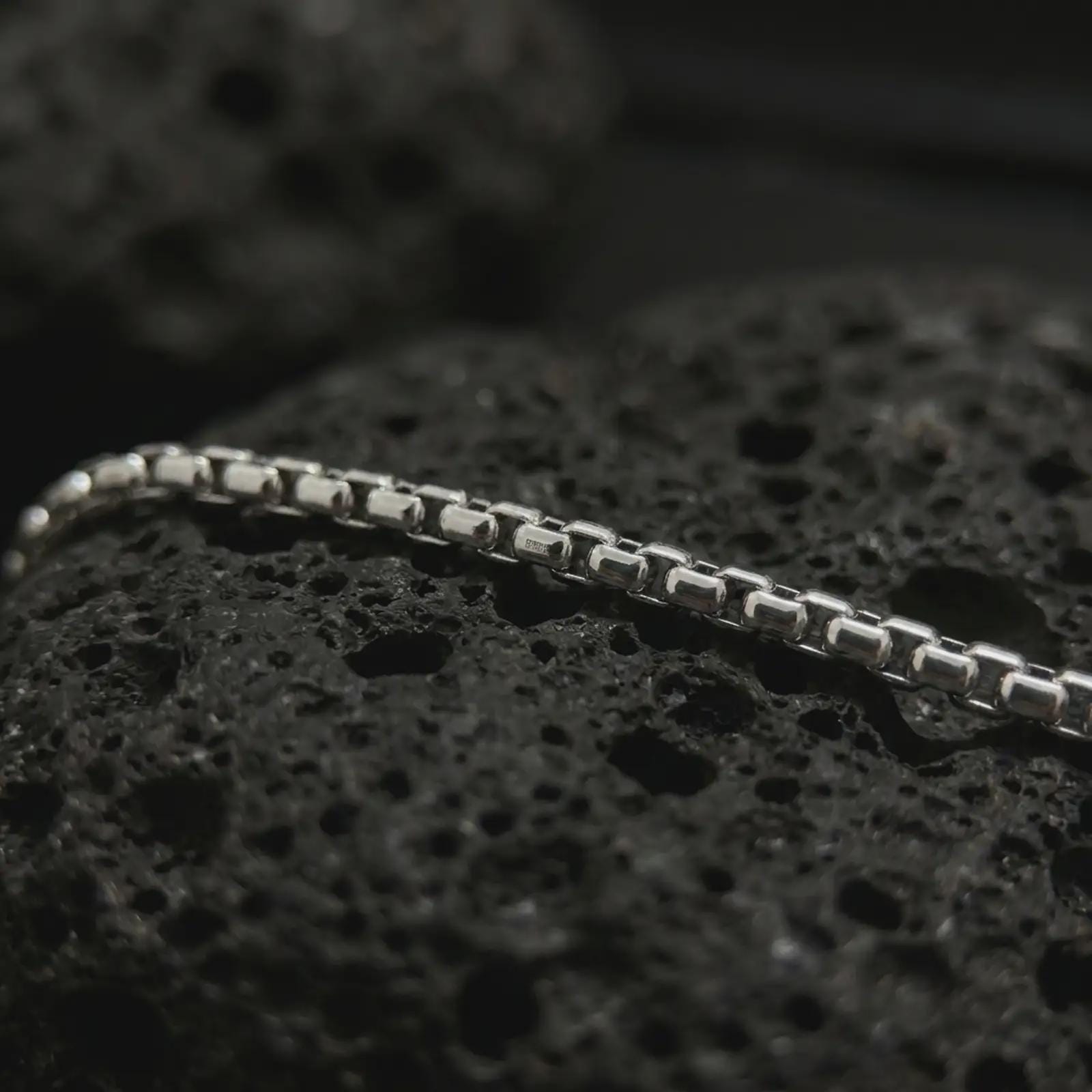 Polished silver box chain necklace on volcanic stone, highlighting the compact geometric links and clean reflective finish.