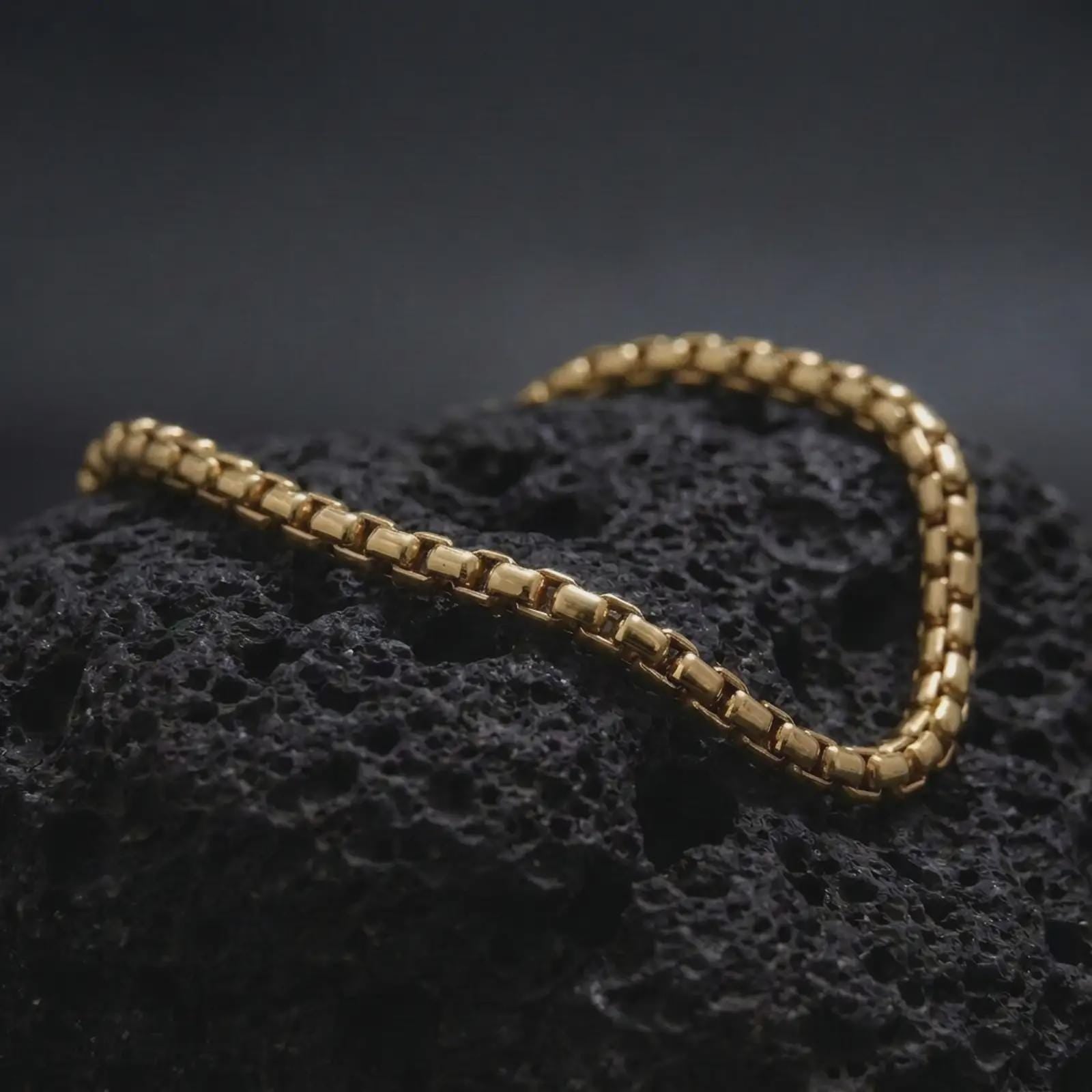 Gold plated box chain bracelet on black volcanic rock, designed with compact rectangular links and a slim masculine profile.