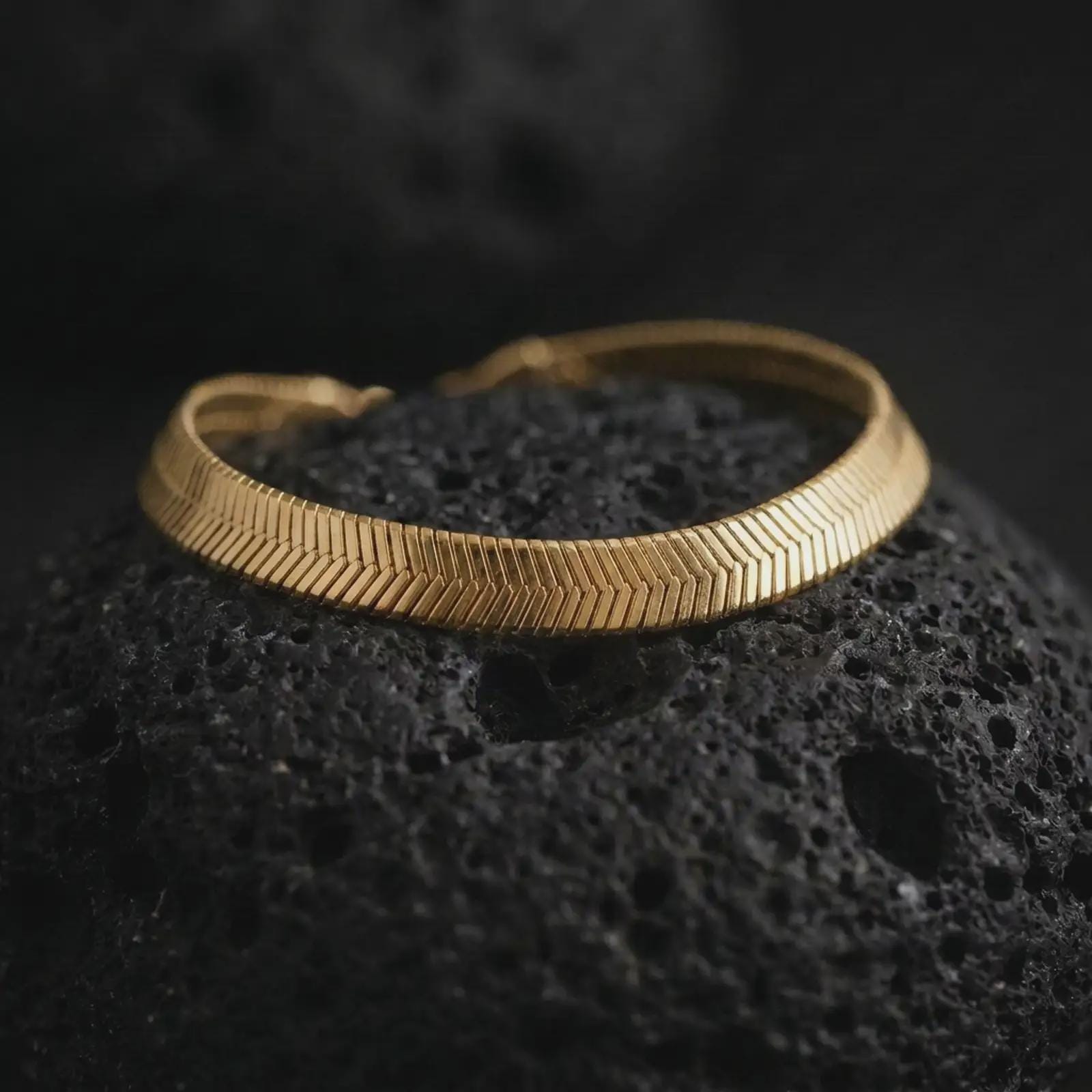 Gold plated flat herringbone bracelet on volcanic rock, highlighting the smooth wrist-hugging shape and structured chevron design.