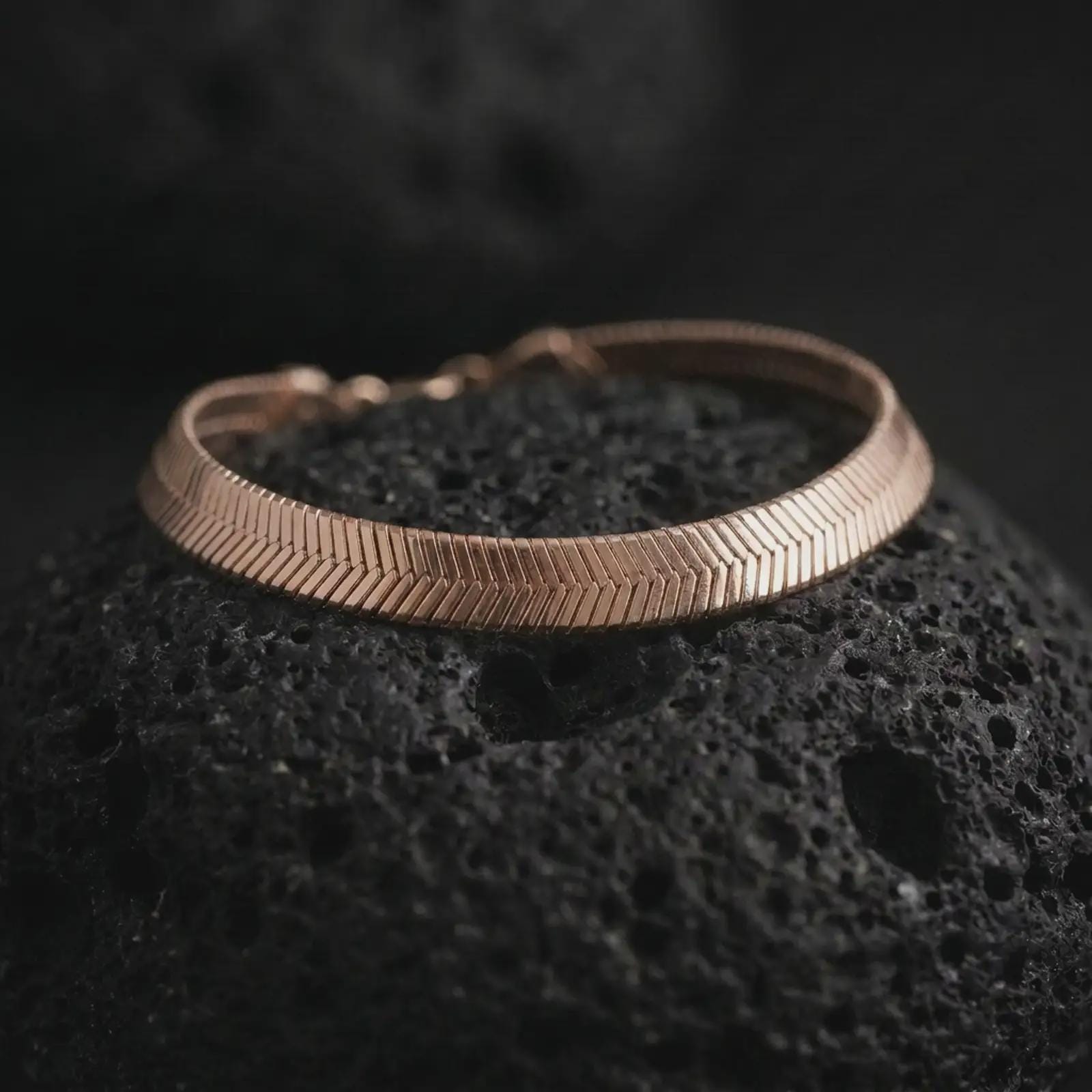 Rose gold plated herringbone bracelet displayed on black lava stone, combining a warm finish with a minimal flat chain silhouette.