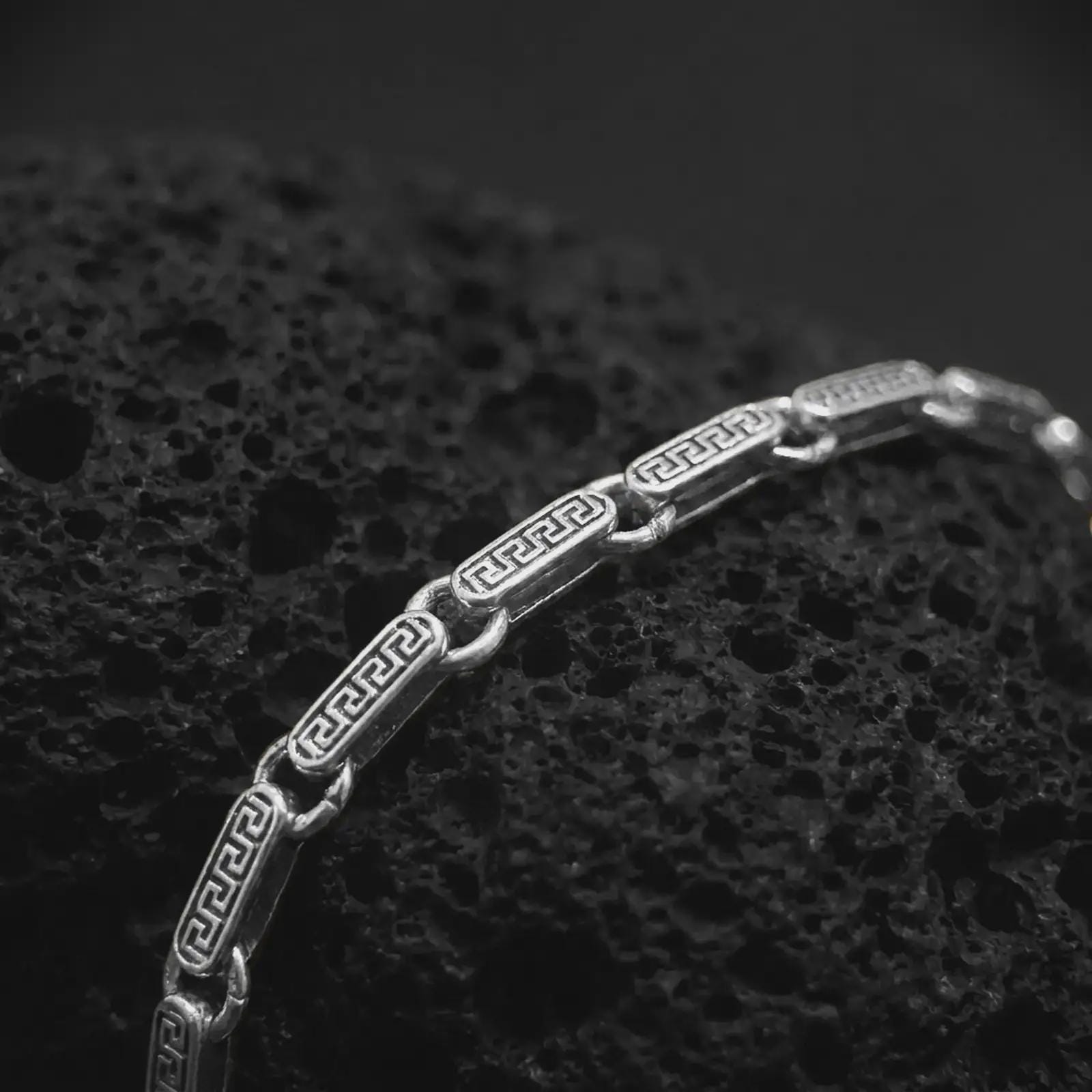 Greek meander chain bracelet in polished silver resting on textured volcanic rock in a detailed macro studio close-up.