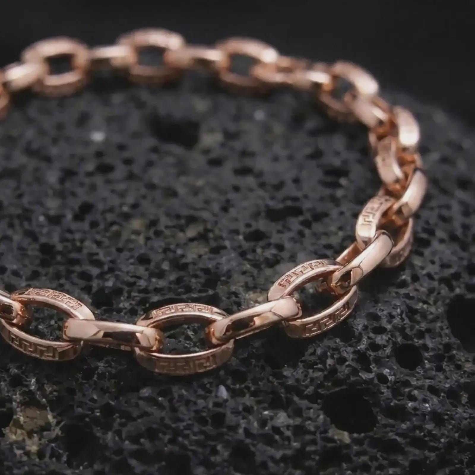 Rose gold plated meander link bracelet arranged on black volcanic rock, featuring repeating Greek key engravings and articulated rectangular connectors in a refined macro close-up.