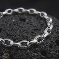 Load image into Gallery viewer, Polished silver Greek meander bracelet displayed across textured black volcanic rock, highlighting the alternating circular and rectangular chain links in a premium macro studio photograph.
