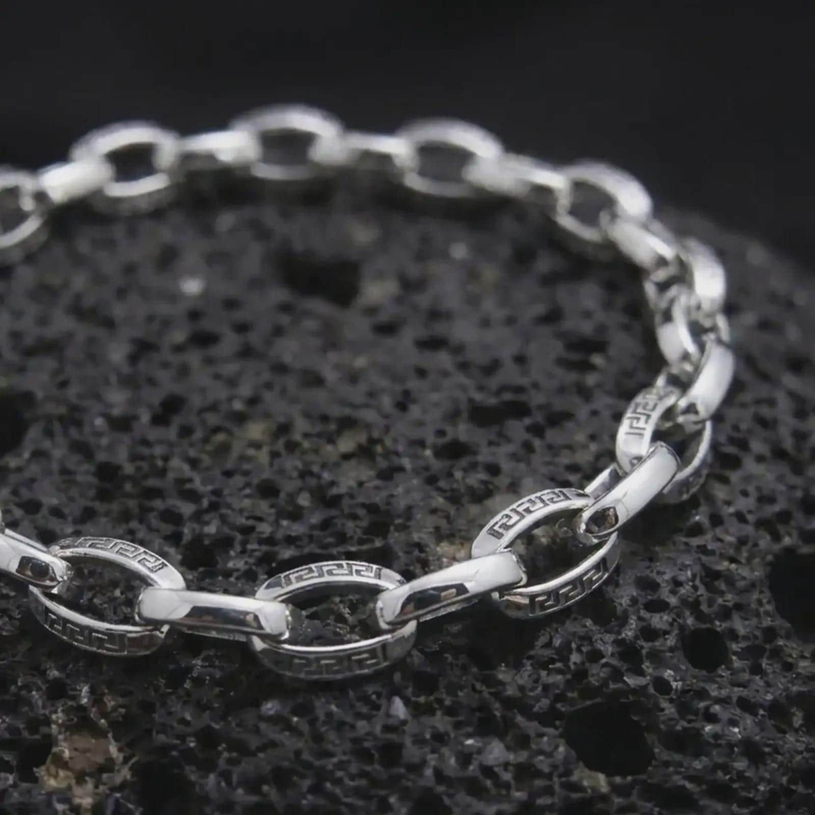 Polished silver Greek meander bracelet displayed across textured black volcanic rock, highlighting the alternating circular and rectangular chain links in a premium macro studio photograph.