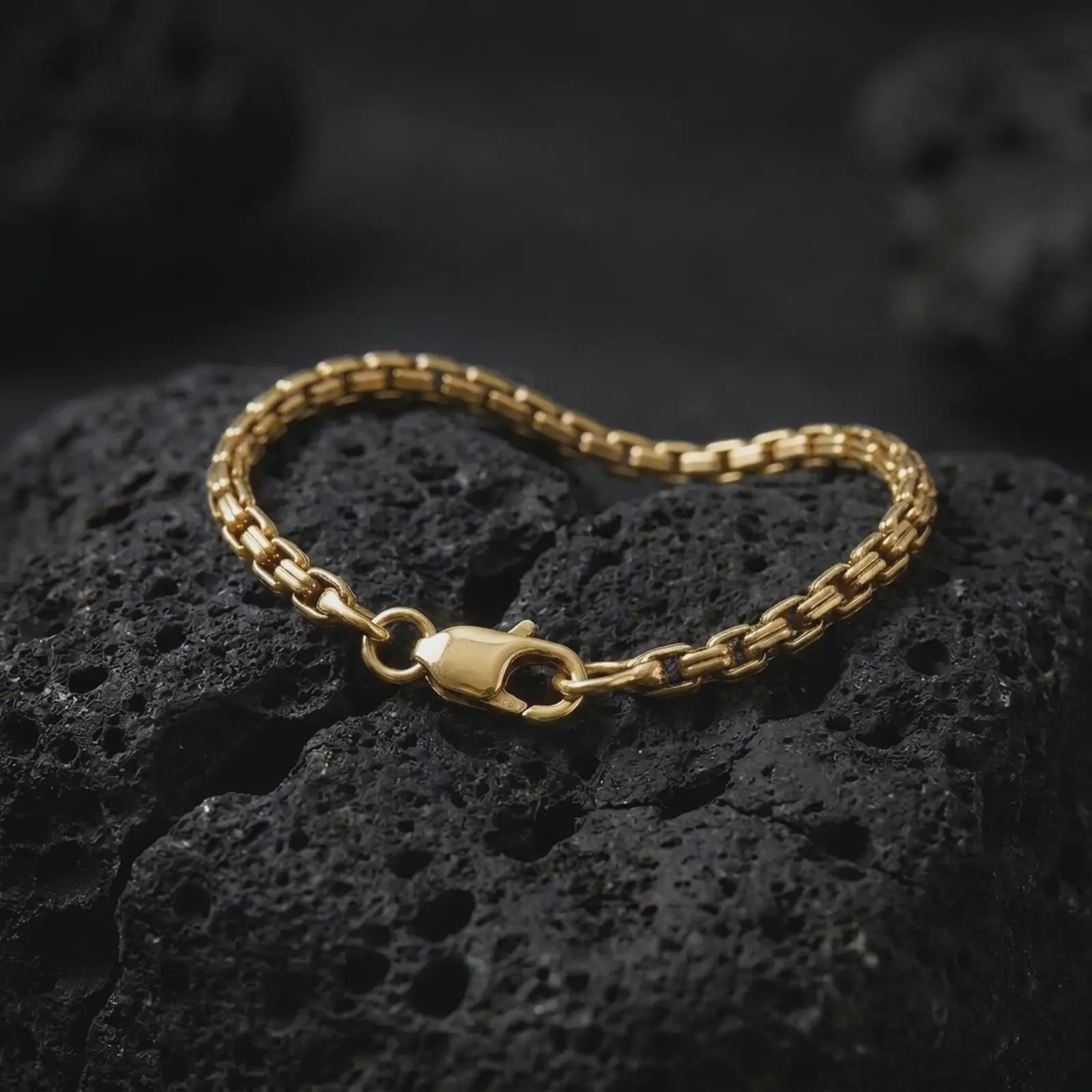 Gold plated men’s box chain bracelet resting across dark volcanic rock, with its narrow geometric link pattern and polished lobster clasp captured in a luxury macro studio close-up.