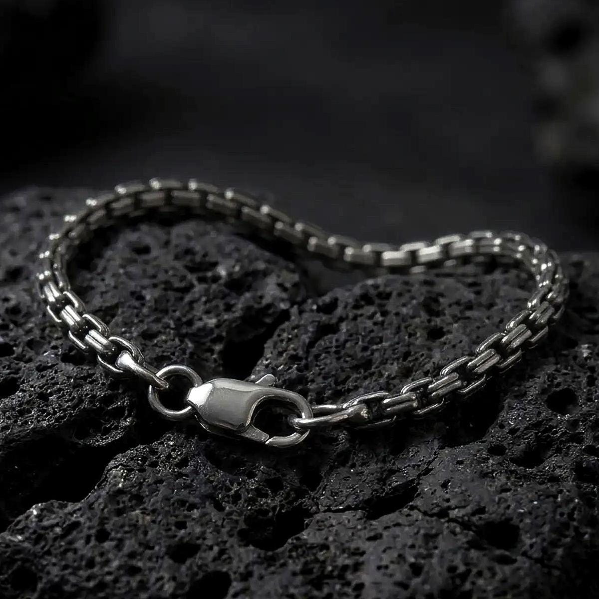 Slim sterling silver box chain bracelet with an oxidized finish arranged in a curved shape on black volcanic rock, showing its compact geometric links and lobster clasp in a macro studio close-up.