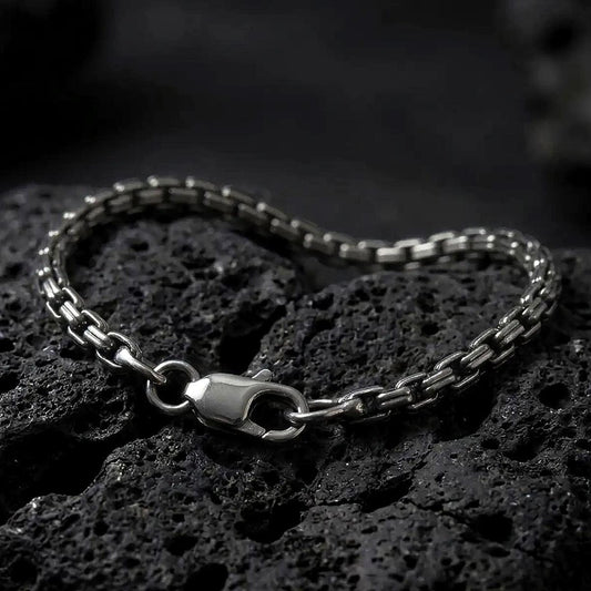 Slim sterling silver box chain bracelet with an oxidized finish arranged in a curved shape on black volcanic rock, showing its compact geometric links and lobster clasp in a macro studio close-up.