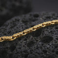 Load image into Gallery viewer, A gold plated Greek key link bracelet lies on rugged volcanic rock, displaying repeating engraved motifs and articulated connecting rings in a luxury macro studio close-up.

