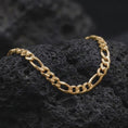 Load image into Gallery viewer, A gold plated 7mm Figaro necklace is displayed on rugged volcanic rock, featuring alternating elongated oval links and compact curb links in a luxury macro studio close-up.
