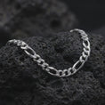 Load image into Gallery viewer, The polished silver 7mm Figaro chain necklace is arranged across textured black volcanic rock, highlighting its traditional 3+1 Italian link pattern in a premium macro photograph.
