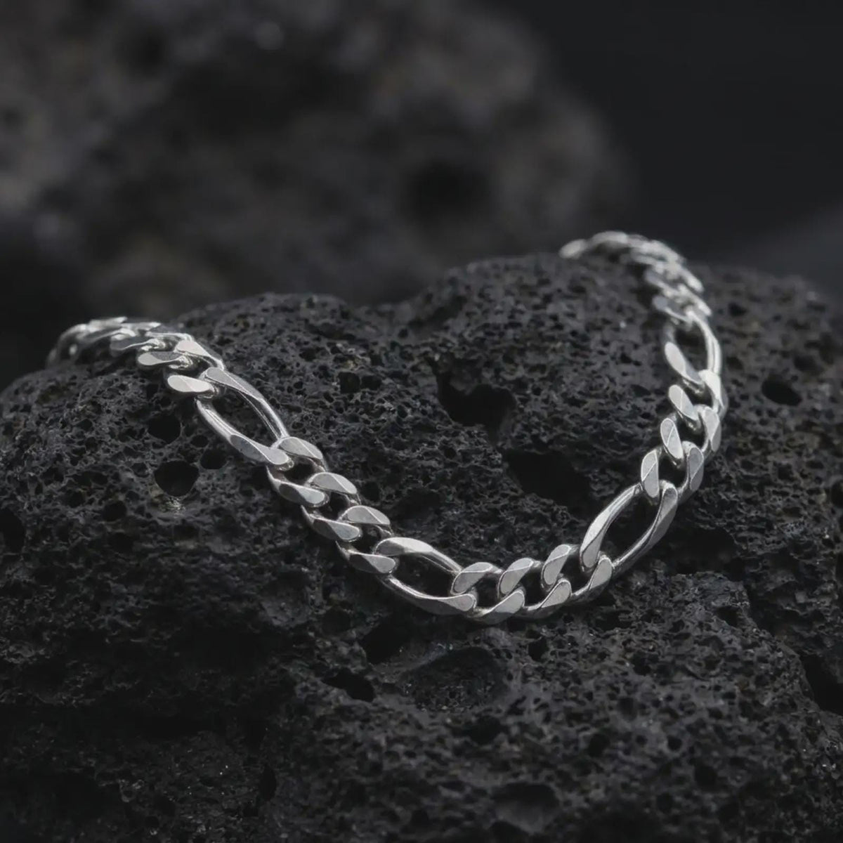 The polished silver 7mm Figaro chain necklace is arranged across textured black volcanic rock, highlighting its traditional 3+1 Italian link pattern in a premium macro photograph.