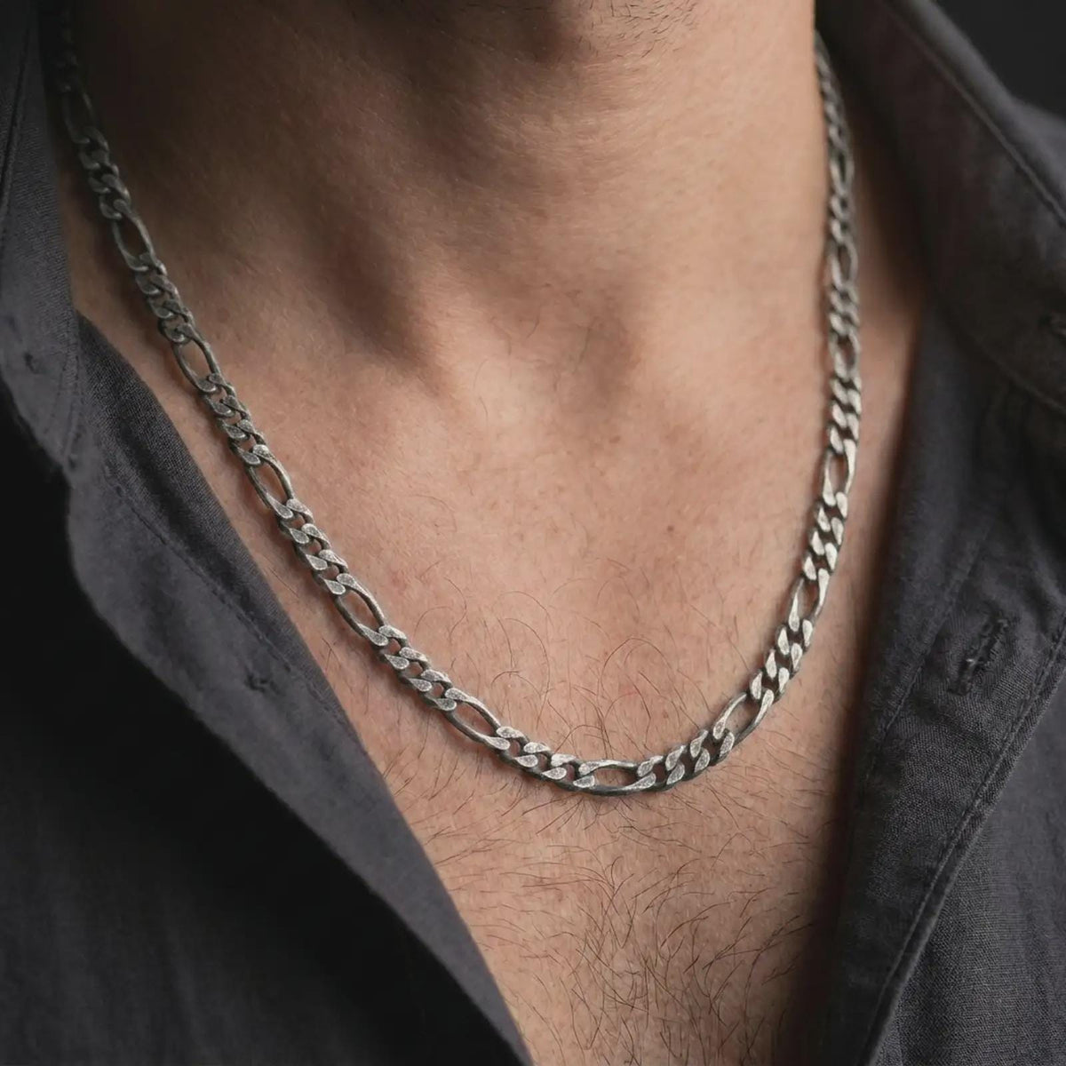 A 7mm Figaro chain necklace with an oxidized finish is worn on a man’s neck, showing the darkened recessed areas and classic Italian link pattern in a masculine lifestyle close-up.