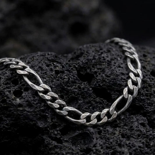 A 7mm Figaro chain necklace with an oxidized finish rests on dark volcanic rock, showing the classic three short links and one elongated oval link in a macro studio close-up.