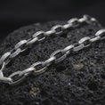 Load image into Gallery viewer, Oxidized Greek key chain necklace rests on dark volcanic rock in a macro studio close-up, showing patterned silver links with ancient-inspired detail.
