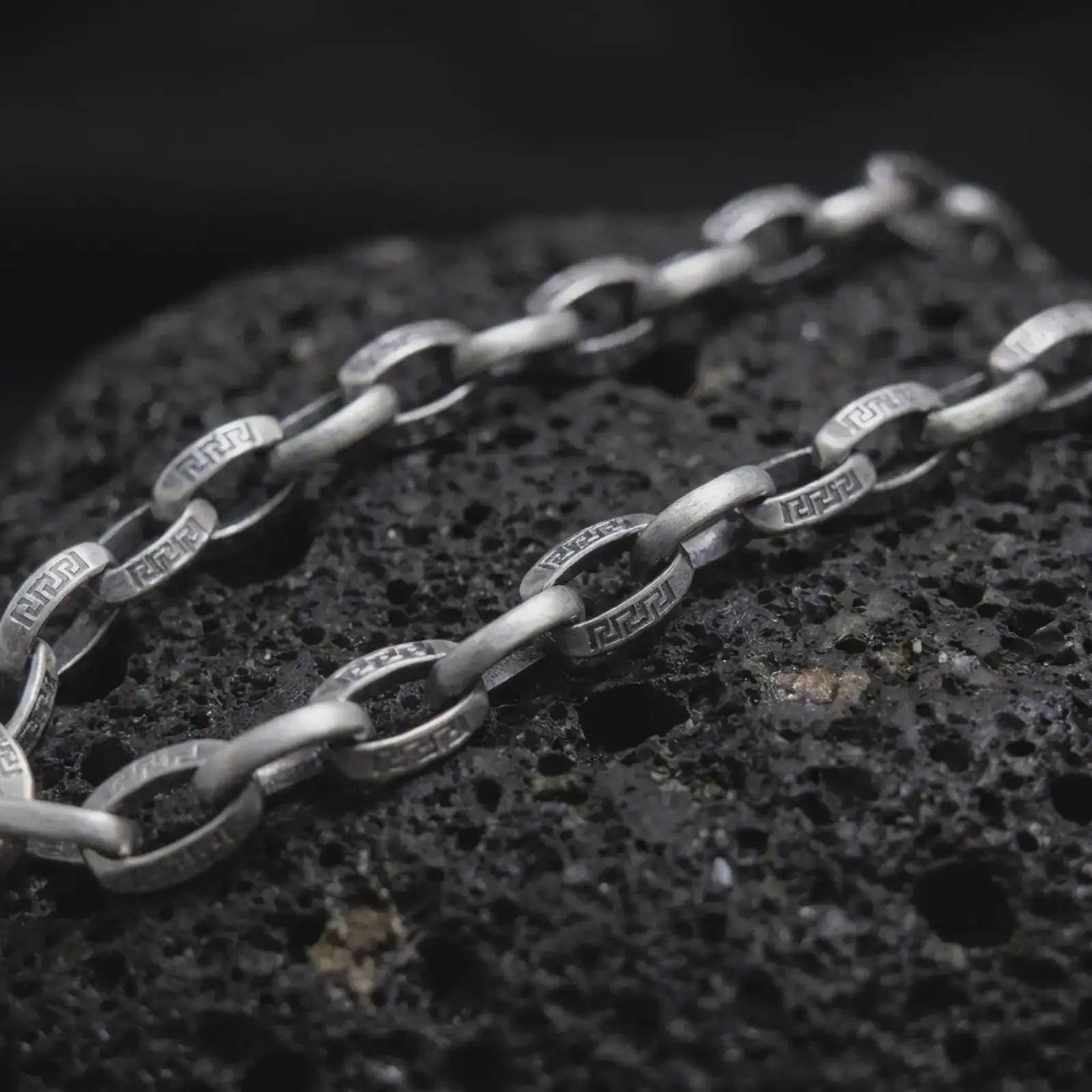 Oxidized Greek key chain necklace rests on dark volcanic rock in a macro studio close-up, showing patterned silver links with ancient-inspired detail.